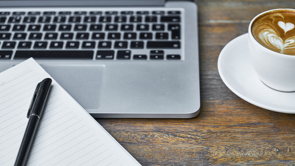 ein Macbook, ein Block mit Stift und ein Cappuccino